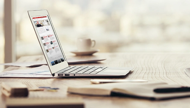 A laptop with web browser open to the SEI website sitting on a desk next to a coffee cup, cell phone, and open book.