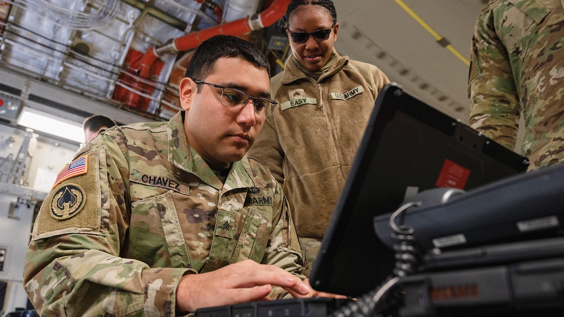 Two US Army service members in uniform work on a laptop inside a facility, with equipment and piping visible overhead.