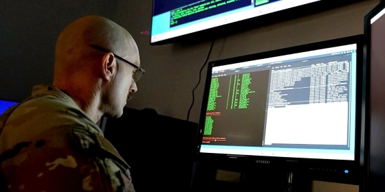 A U.S. Airman analyzing code on multiple monitors in a dark room.