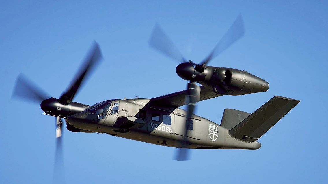 Tiltrotor aircraft in flight with rotating propellers, shown against a clear blue sky from a side view.