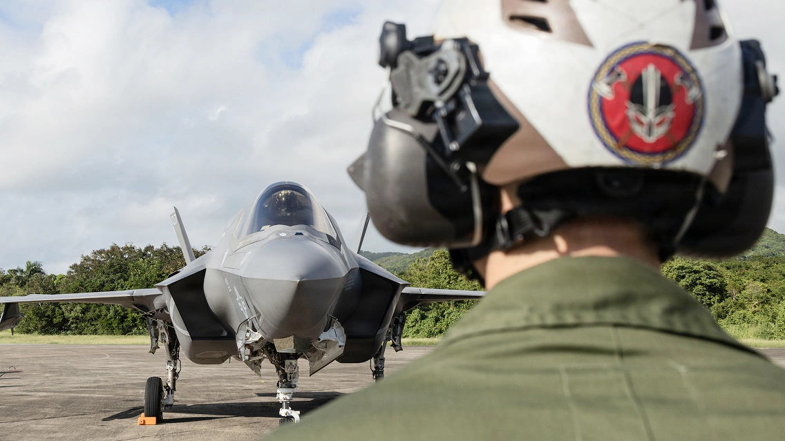 Service member wearing a helmet faces a parked F-35 jet on a runway, with the aircraft centered against a cloudy sky.