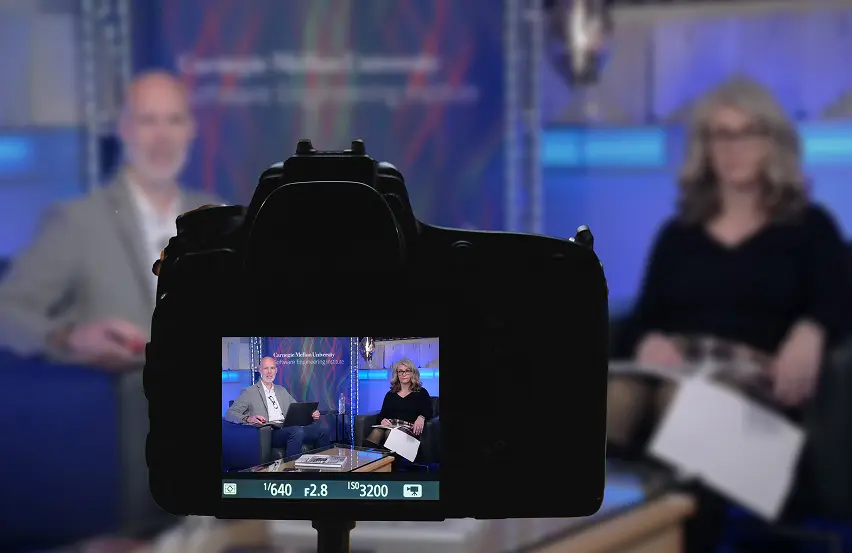 Close-up of a DSLR camera's LCD viewfinder, showing a still frame from a podcast recording session. The figures in the episode are blurred in the background.